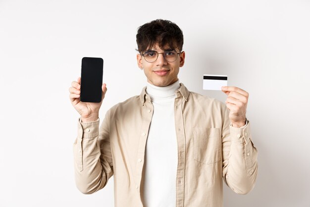Online shopping young modern guy showing plastic credit card and empty smartphone screen demonstrate...