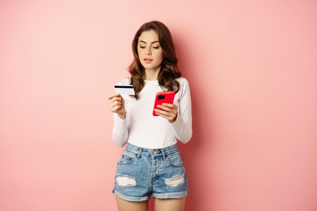 オンラインショッピング。若い女性はオンラインで注文し、アプリで購入し、スマートフォンとクレジットカードを持って、ピンクの背景に立っています