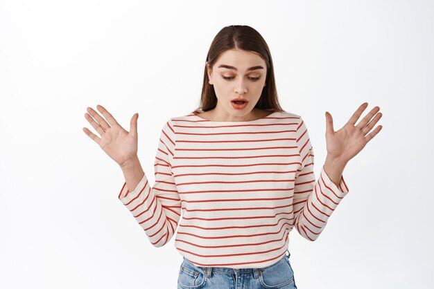 Oops dropped something Gasping girl raise hands sideways and looks down surprised see something bellow standing ambushed against white background