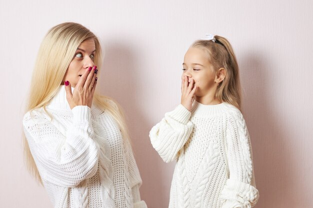 Oops, Omg. Adorable little girl and her young mother both in white sweaters posing isolated keeping hands on mouths, being astonished with big sale prices, going shopping to buy Christmas presents