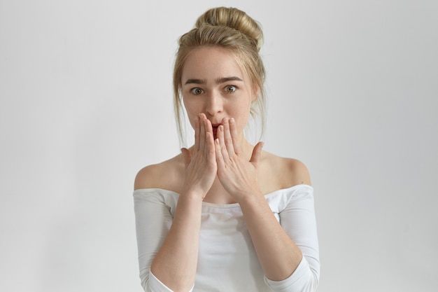 Oops Picture of emotional beautiful young lady with hear bun covering mouth with hands, trying not to tell secret, being surprised with astonishing news or gossip, staring with shocked look