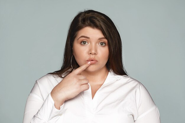 Oops. Picture of glamorous attractive obese young dark haired woman in white shirt touching lip and looking with clueless expression