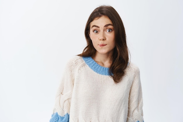 Oops she did small mistake Silly cute girl biting lip and looking innocent at camera raising eyebrows with clueless face standing against white background in sweater