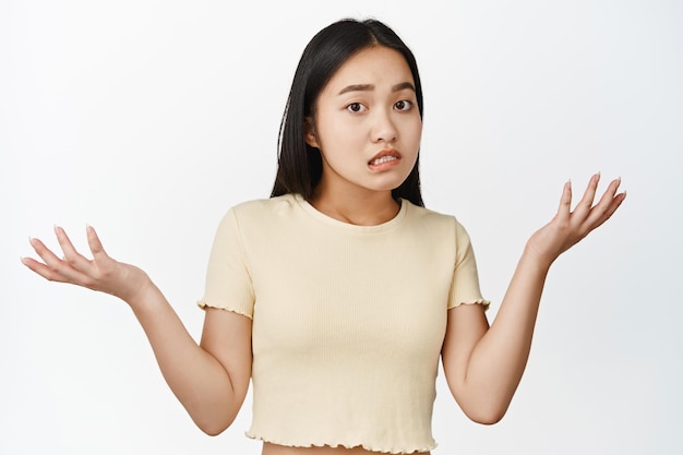 Oops sorry Apologizing asian girl spread hands sideways shrugging shoulders and looking puzzled dont understand standing confused against white background