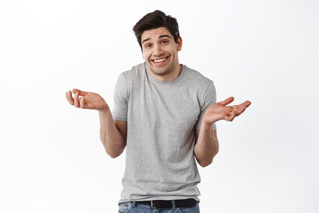 Oops sorry, my mistake. Man shrugging and smiling awkward, feeling guilty, apologizing and looking coy at camera, standing over white background