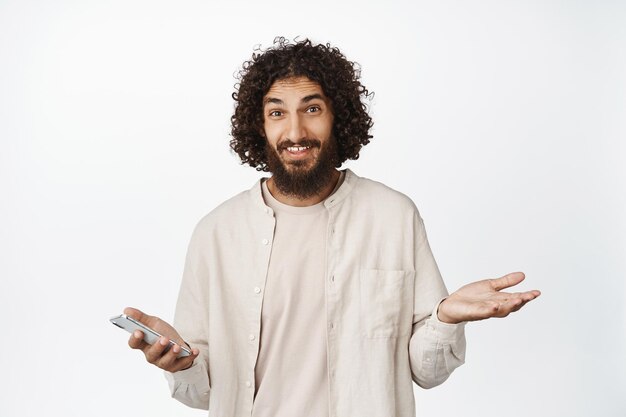 Oops sorry Smiling guilty guy shrugging shoulders and holding smartphone standing over white background