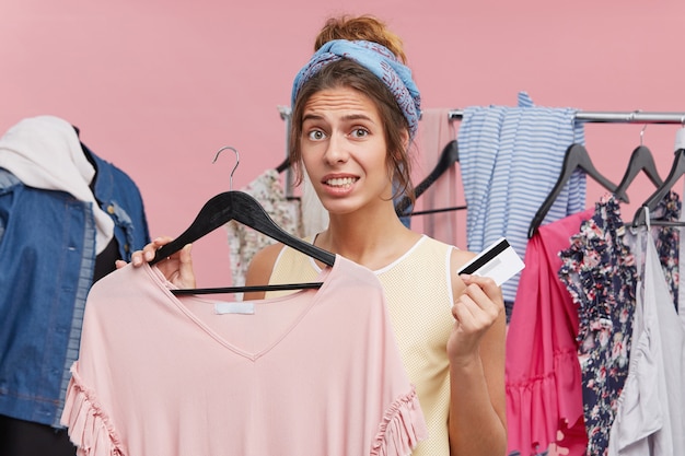 Free photo oops! worried woman holding dress in boutique in one hand and credit card in other, feeling puzzled that she hasn`t money on her account for paying her purchase. unforeseen expanses on clothes