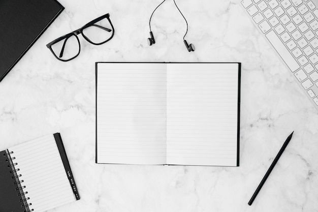 An open blank notebook with eyeglasses; diary; pencil; earphone; keyboard on white desk