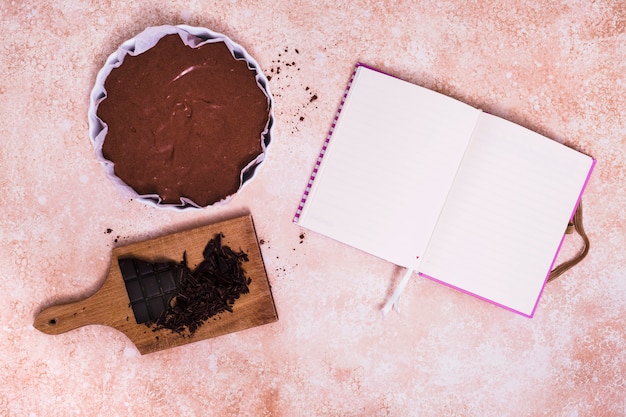 Free photo an open blank white diary with cake and broken chocolate bar on chopping board over the textured background