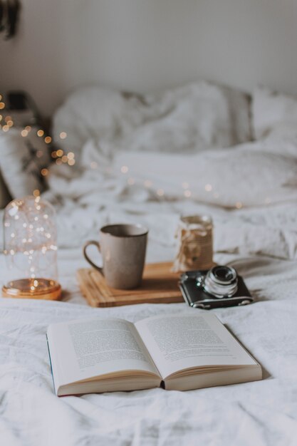 Open book, a camera, a tray and a mug on a bed with white sheets