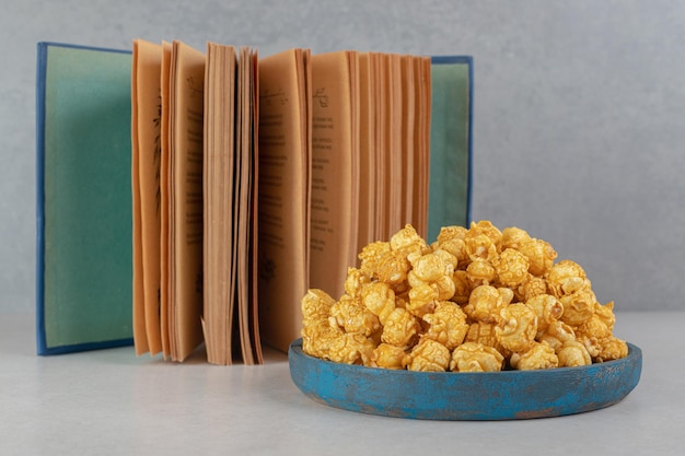 Free photo open book next to an overfilled small tray of caramel popcorn on marble table.