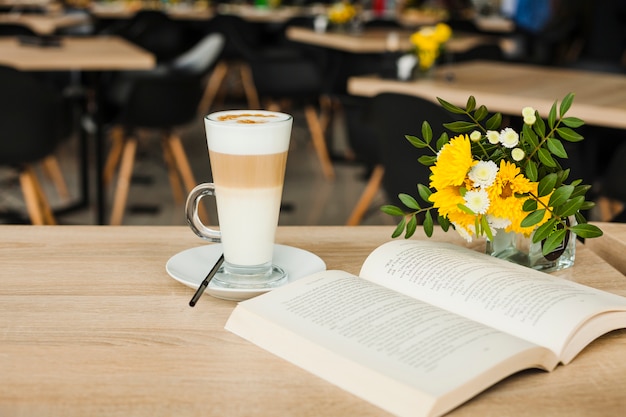 Открытая книга с чашкой кофе латте и вазой из свежих цветов над деревянным столом