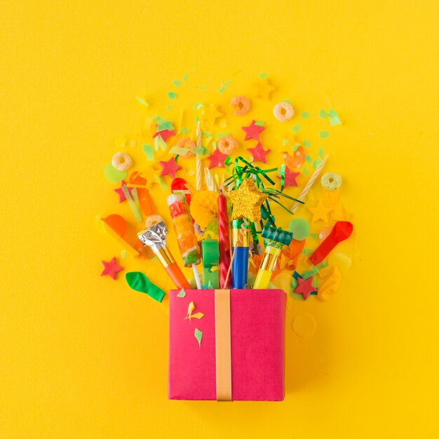 Open gift box with candies and party accessories on yellow background