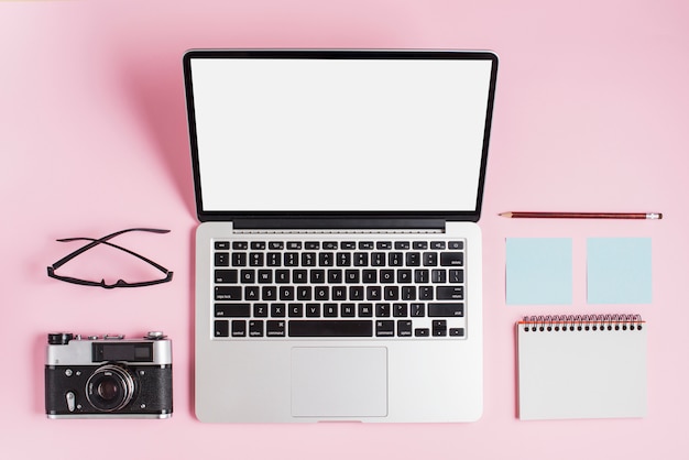 Открытый ноутбук; камера; очки; карандаш и спиральный блокнот на розовом фоне