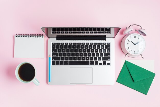 An open laptop; pencil; alarm clock; spiral notepad and envelope on pink background