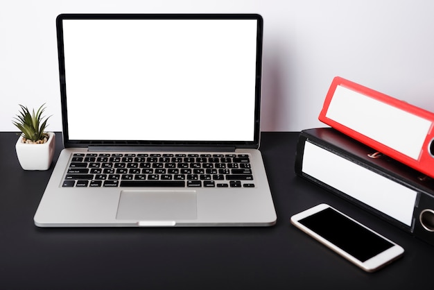 An open laptop with blank white screen; cell phone and paper clips on black desk against white wall