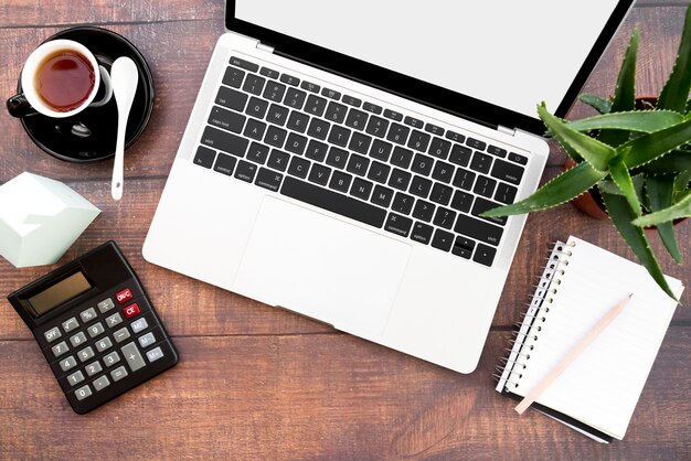 An open laptop with coffee cup; spiral notebook; calculator; paper house model and aloe vera plants on wooden table