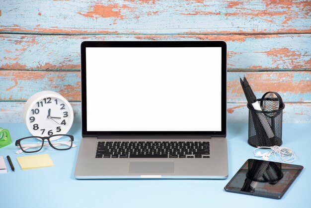 Открытый ноутбук с белым пустым экраном; цифровой планшет и канцелярские принадлежности на синем столе