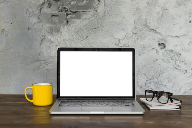 Open laptop; yellow coffee mug; spectacle; and diary on wooden table with background of weathered wall