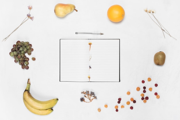 An open notebook and pen surrounded with different fruits over white backdrop