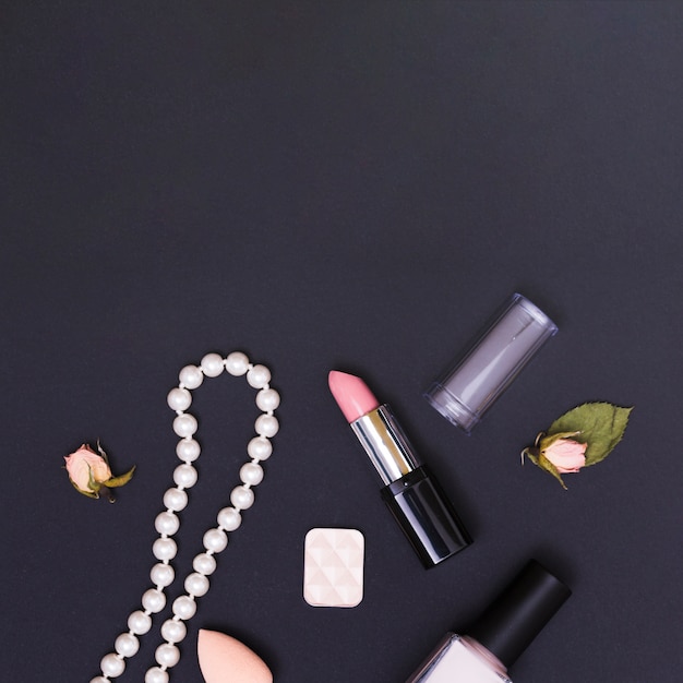 Free photo an open pink lipstick; nail polish; necklace; sponge; rose bud and eyeshadow on black background