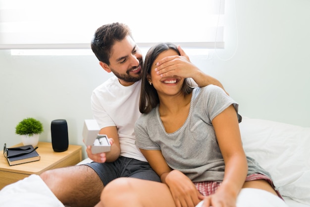 Free photo don't open your eyes. romantic man in love covering his girlfriend's eyes to surprise her with an engagement ring after waking up together in bed