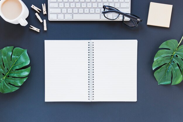 Opened notebook near keyboard and coffee cup on desk with leaves and glasses