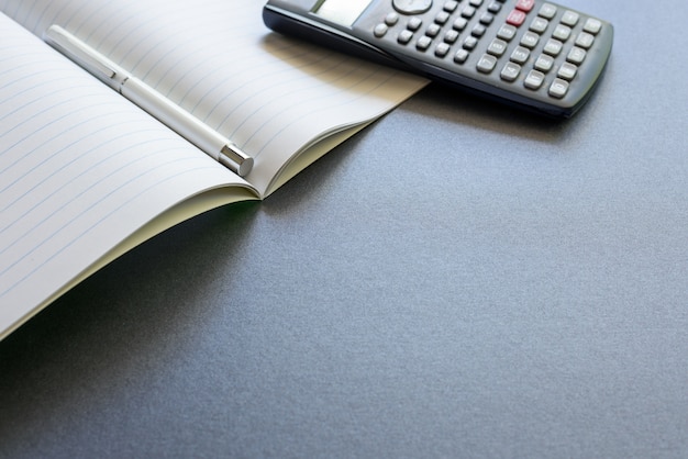 Un notebook aperto con una penna e una calcolatrice, su sfondo grigio scuro, scuola di scena o ufficio.