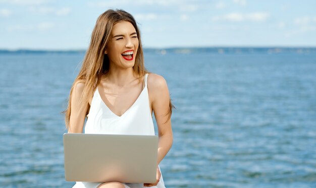 ラップトップストリップを持って公然と笑っている女性は、湖畔に向かって脇に置きます。高品質の写真