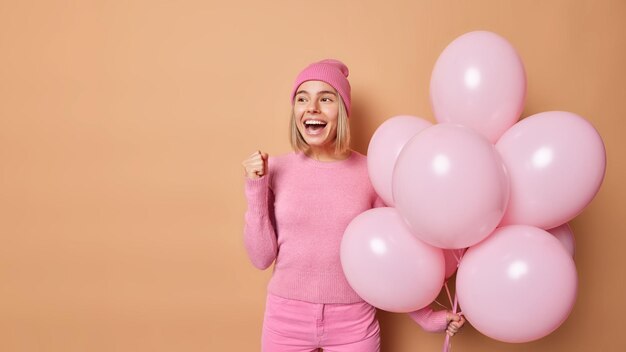 Opimistic joyful European girl clenches fist and exclaims happily hears good news wears pink hat jumper and trousers holds helium balloons isolated over brown background blank copy space away