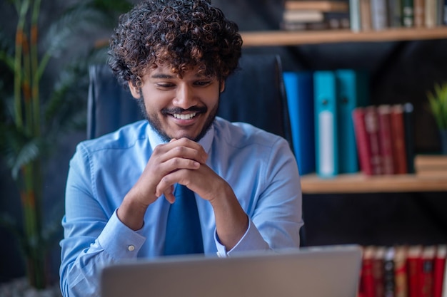 Optimism. Bearded young man with dark curly hair holding his hands near beard sitting in office at table smiling looking at laptop screen