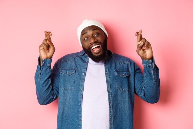 Optimistic african-american man making wish, holding fingers crossed and smiling, standing over pink background