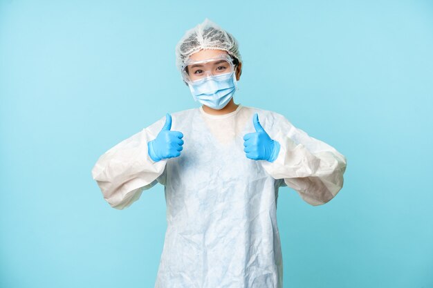 낙관적인 아시아 간호사나 의사는 개인 보호 장비를 착용하고 엄지손가락을 치켜들고 코로나바이러스 예방 캠페인, 파란색 배경을 보여줍니다.