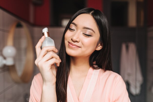 Optimistic Asian woman smiles with closed eyes and holds jar of cream