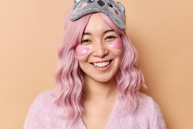 Оптимистичная азиатка с крашеными розовыми волосами счастливо улыбается, накладывает гидрогелевые пластыри под глаза, чтобы уменьшить морщины, носит повседневный джемпер и маску для сна, изолированные на коричневом фоне. Время красоты