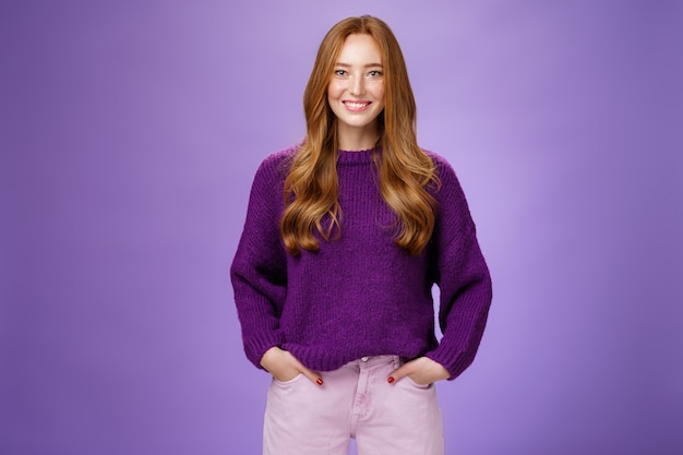 Оптимистичная привлекательная рыжая девушка в 20-х годах в фиолетовом свитере стоит наготове и заряжена энергией на фиолетовом фоне, дружелюбно и уверенно улыбаясь в камеру, демонстрируя готовность повеселиться.