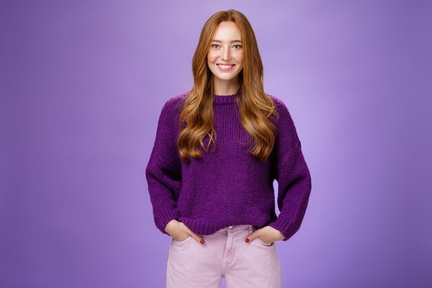 Optimistic attractive ginger girl in 20s wearing purple sweater standing ready and energized over violer background smiling friendly and confident at camera, showing readiness to have fun.