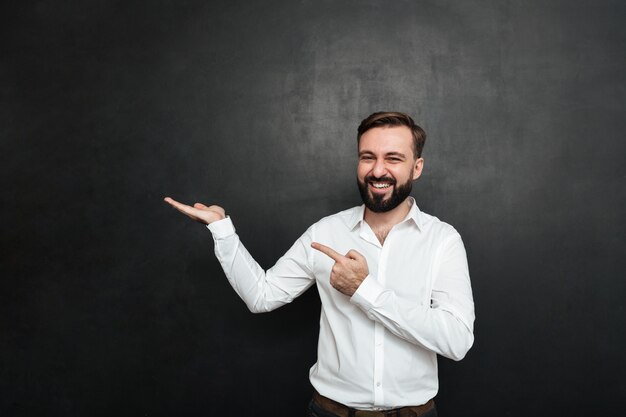 Optimistic bearded man pointing index finger while holding thing on palm, demonstrating or advertising over dark gray copy space