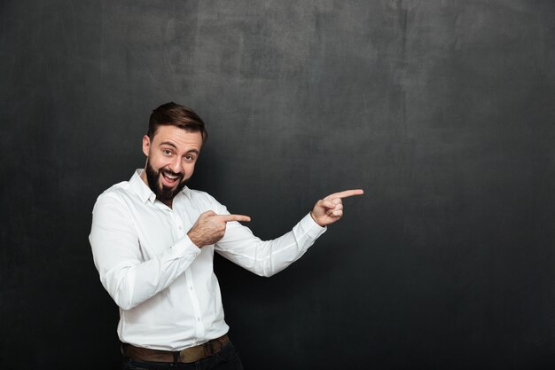 Optimistic bearded man in white shirt pointing index fingers aside, demonstrating or advertising over dark gray copy space