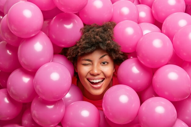 Optimistic birthday woman has fun and winks eye smiles joyfully poses against many pink balloons