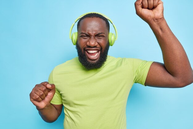 Оптимистичный беззаботный бородатый мужчина танцует в ритме музыки, одетый в зеленую футболку, слушает музыку, изолированную над синей стеной