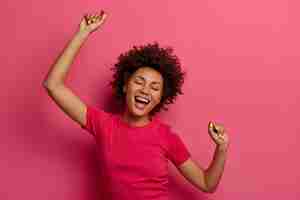 Free photo optimistic carefree curly young woman raises arms and dances to favorite awesome music, moves with rhythm of song, wears casual t-shirt, isolated over pink wall, forgets about all troubles