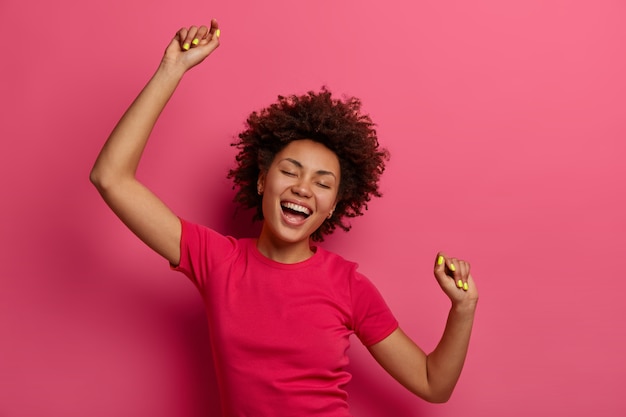Optimistic carefree curly young woman raises arms and dances to favorite awesome music, moves with rhythm of song, wears casual t-shirt, isolated over pink wall, forgets about all troubles