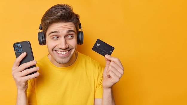 Бесплатное фото Оптимистичный беззаботный красивый мужчина держит современный смартфон, а банковская карта использует приложение на мобильном телефоне для онлайн-платежей, изолированных на желтом фоне, пустое место для вашего рекламного контента