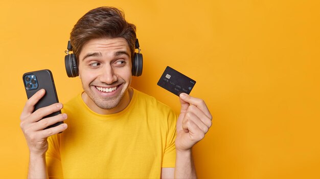 楽観的な屈託のないハンサムな男は、現代のスマートフォンを保持し、銀行カードは、プロモーションコンテンツの黄色の背景の空白スペースに分離されたオンライン支払いのために携帯電話のアプリケーションを使用します