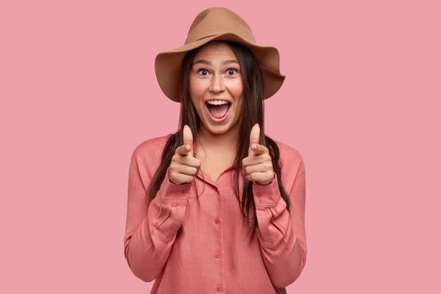 Optimistic Caucasian woman with dark hair, freckled skin, makes face and points with finger gun gesture, smiles broadly