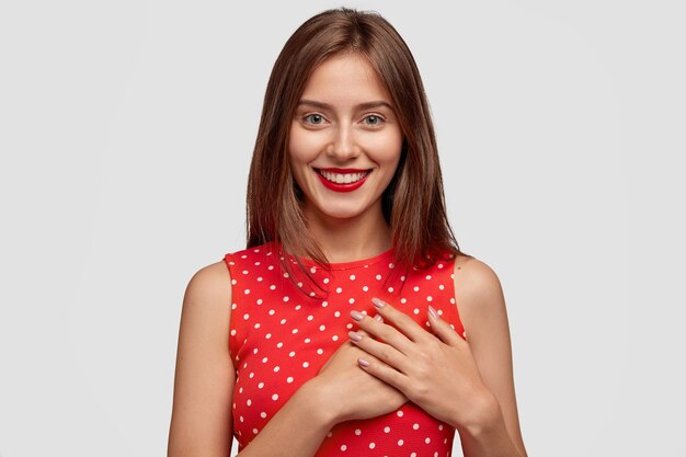 暖かい言葉に感謝している楽観的な陽気なブルネットの女性は、胸に両手を保ち、赤い口紅を持っています
