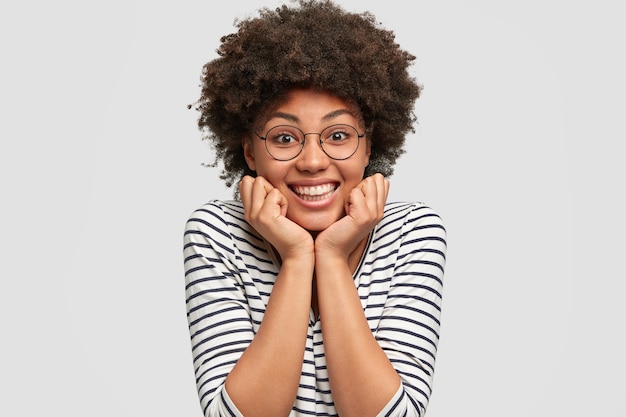 La femmina afroamericana riccia ottimista ha un sorriso a trentadue denti, tiene le mani sotto il mento, ascolta la piacevole storia di un amico, indossa abiti casual e occhiali rotondi, isolato su un muro bianco.