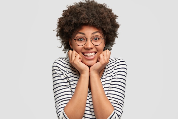 Optimistic curly African American female has toothy smile, keeps hands under chin, listens pleasant story from friend, wears casual clothes and round glasses, isolated over white wall.