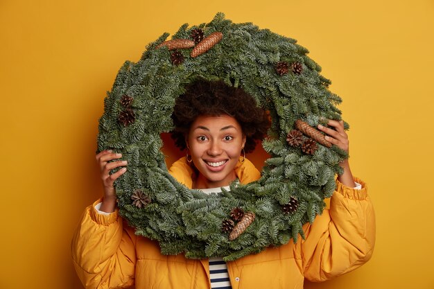 Optimistic curly woman looks through handmade Christmas wreath, being in good mood, wears down padded coat, stands against yellow background.